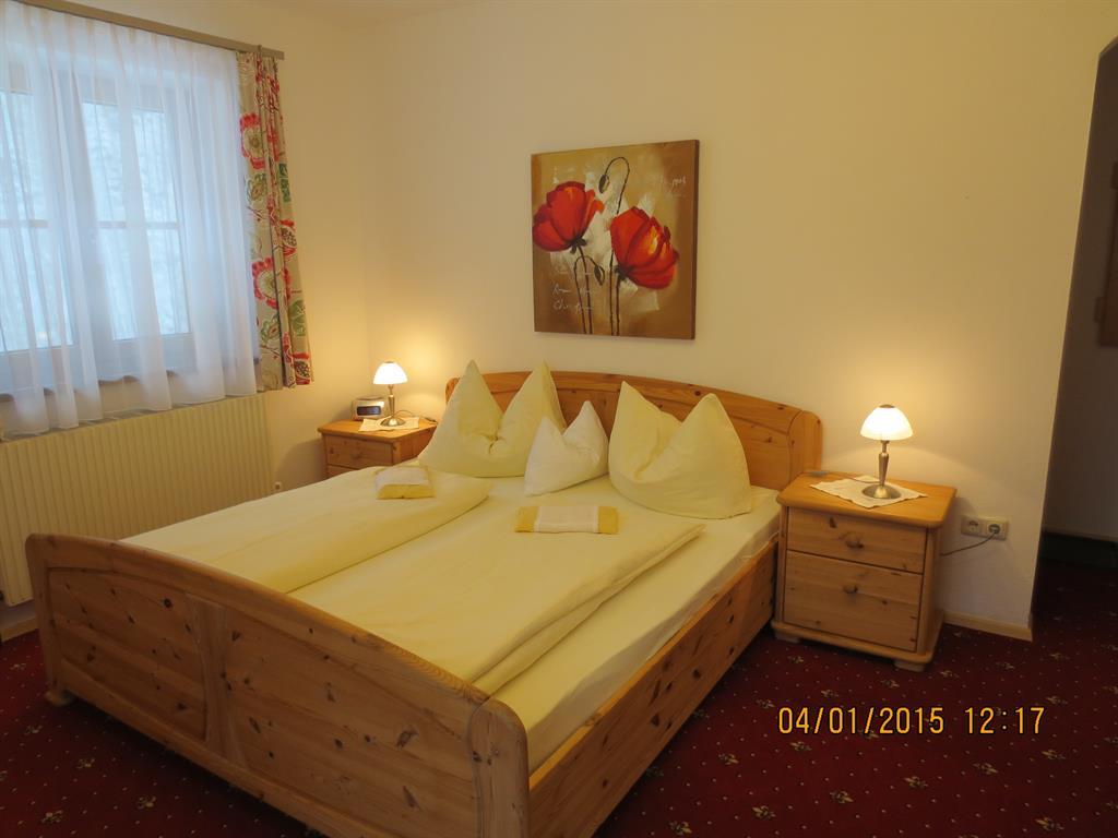 A cozy bedroom with a large double bed and elegant cushions. The walls are light and there are modern lamps as well as a decorative picture with red flowers.