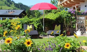A beautiful garden with colorful flowers and two lounge chairs under a red sun umbrella. In the background, mountains and a house are visible.