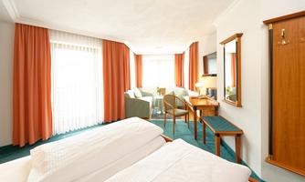 A bright hotel room with two single beds, a sofa, and a table. The windows are fitted with curtains and the room is well furnished.