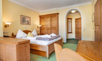 A cozy room with a double bed and wooden furniture. The walls are in a warm yellow tone and there are natural light sources.