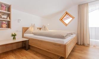 A cozy bedroom with a double bed and wooden floor. There is a window with a small table and a flower decoration.