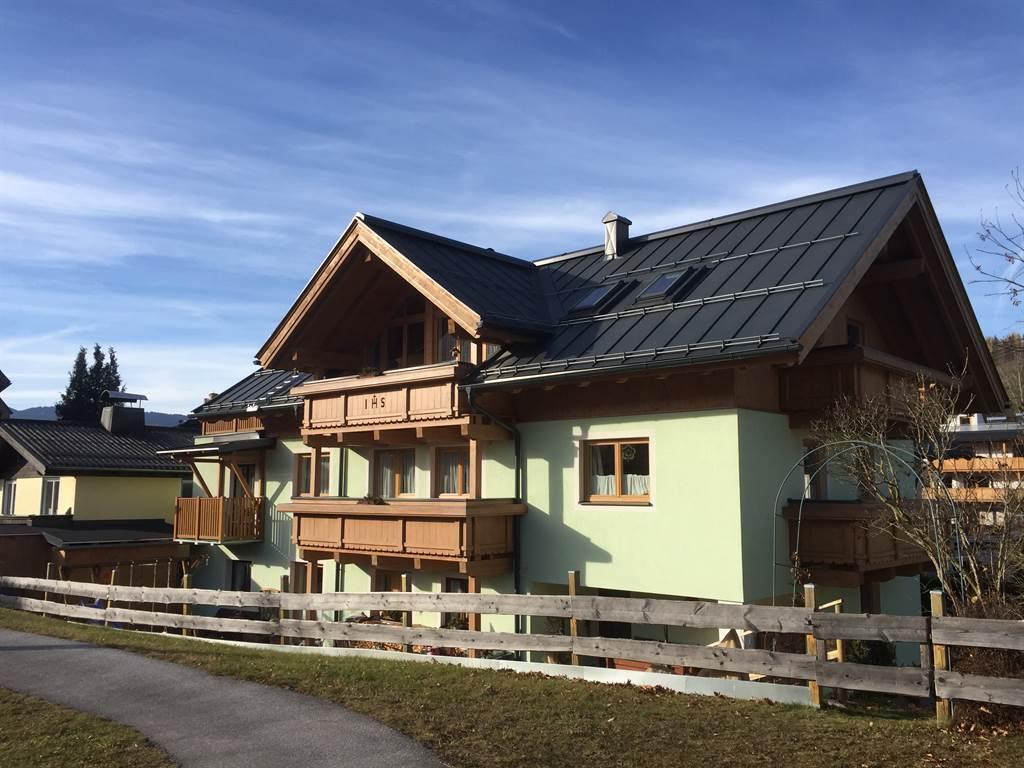 A modern house in Alpine style with a green-painted ground floor and wooden decorations. The surroundings are framed by a tranquil landscape and a paved path.