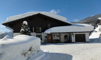 A cozy wooden house with a snow-covered roof. The surroundings are wintry with mountains in the background and a clear blue sky.