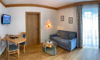 A cozy living space with a seating area, a table, and a sofa. The room has bright wooden walls and a balcony with curtains.