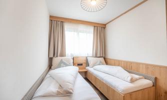 A modern bedroom with two single beds and light wood interiors. Large windows let in a lot of daylight.