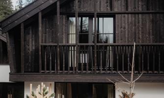 A modern wooden house with a slanted roof and balcony. In the foreground, plants and bushes are growing.