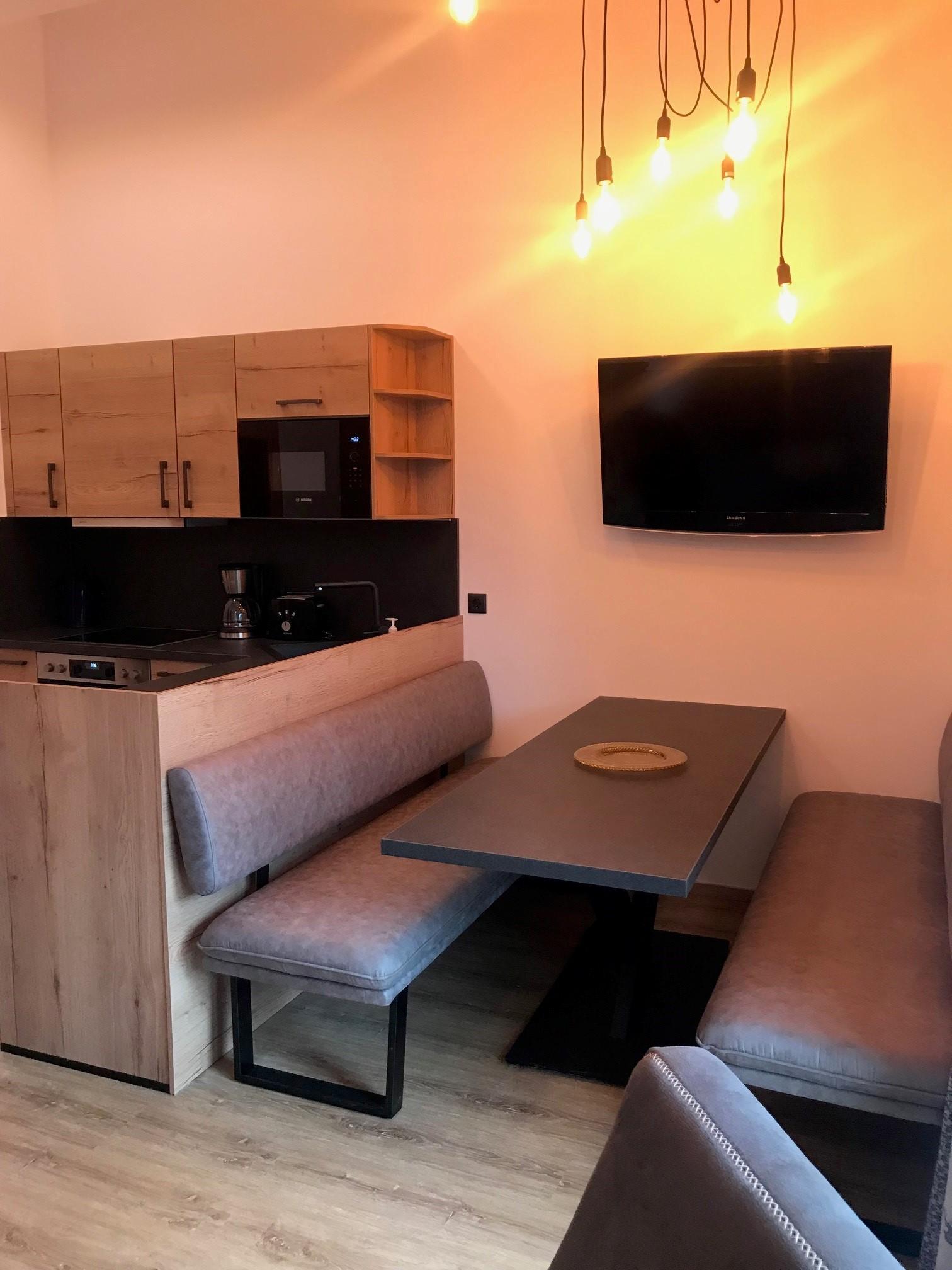 A modern kitchen with a dining area and a television. The decor is simple and in warm wood tones.