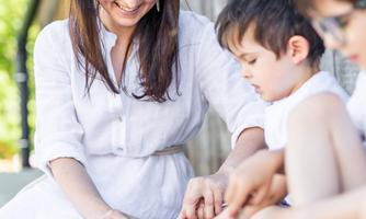 A smiling woman is sitting outside with two children and sharing food. The background is a wooden wall and it is sunny.