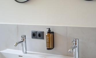 A modern bathroom with two sinks and a large mirror shaped like sunglasses. The wall is light, and the fixtures are made of shiny stainless steel.