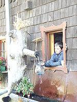 A boy is looking out of a window and holding a water container in his hand. Water is flowing from an old log next to the window.