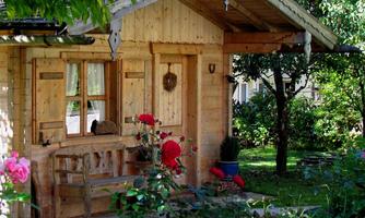 A cozy wooden house stands in a garden with colorful flowers. The house has a porch and is surrounded by green trees.