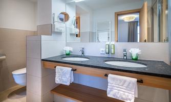A modern bathroom with two sinks and an elegant wooden shelf. The space is bright and well-lit, with minimalist furnishings.