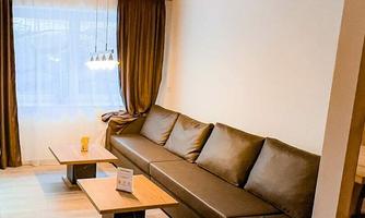 A modern furnished living space with a large brown couch and light wood details. There is a small table and large windows with curtains.