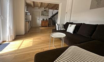 A cozy living room with a dark sofa and a small table. In the background, there is an open kitchen with wooden flooring and plenty of natural light.
