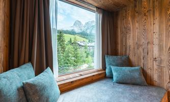 A cozy window seat with soft cushions provides a view of green meadows and mountains. The wooden walls give the room a warm atmosphere.
