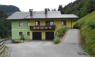 A beautiful two-story house in green and yellow. It is located in a rural setting, surrounded by mountains and trees.