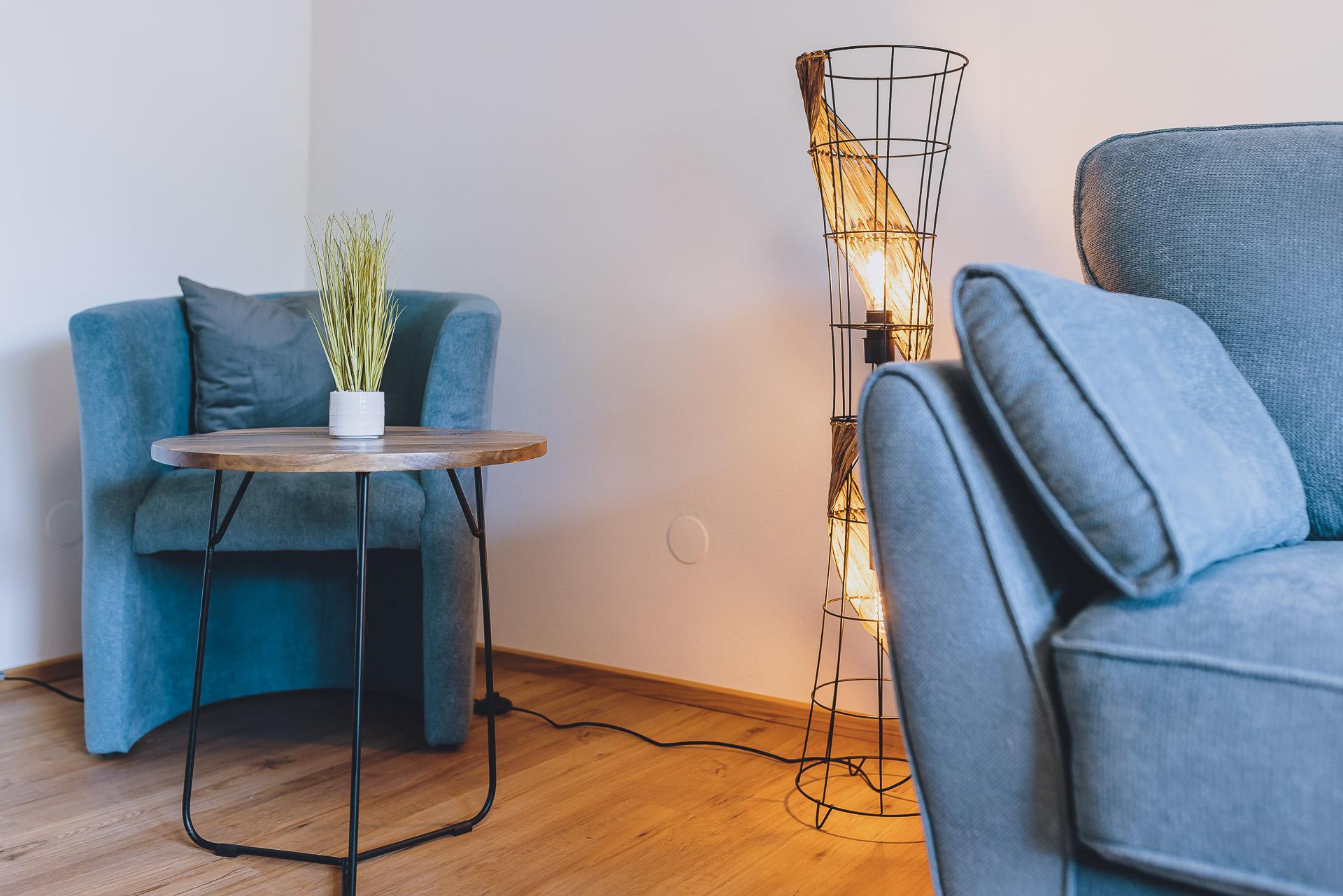 A stylish living room with a blue armchair, a round table, and a modern lamp. The wooden floor complements the cozy atmosphere.