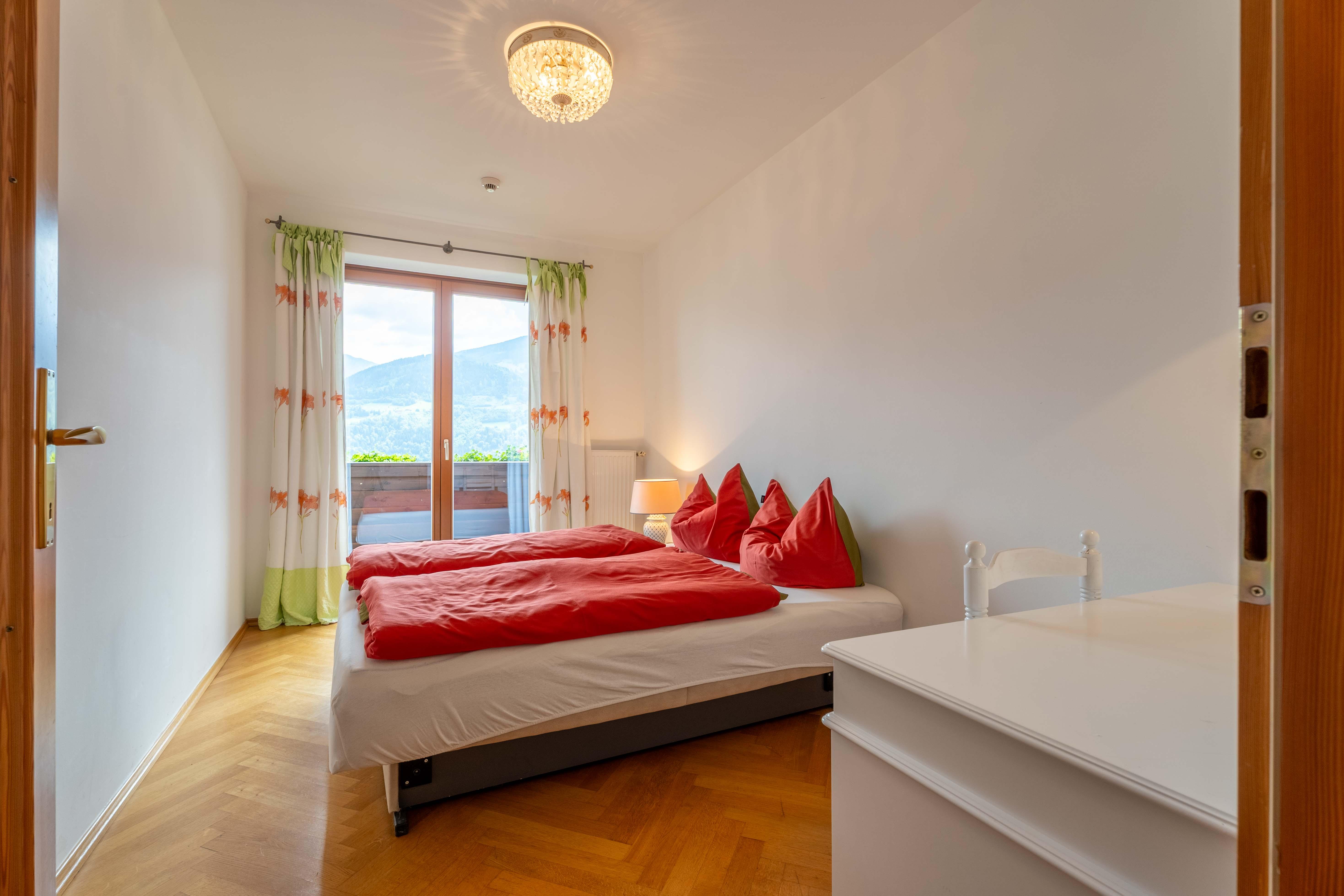 A bright bedroom with a double bed and red pillows. Large windows offer a view of the mountains and bring in plenty of daylight.