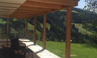 A cozy terrace with a wooden railing and modern chairs. In the background, green hills and forests stretch under a clear sky.