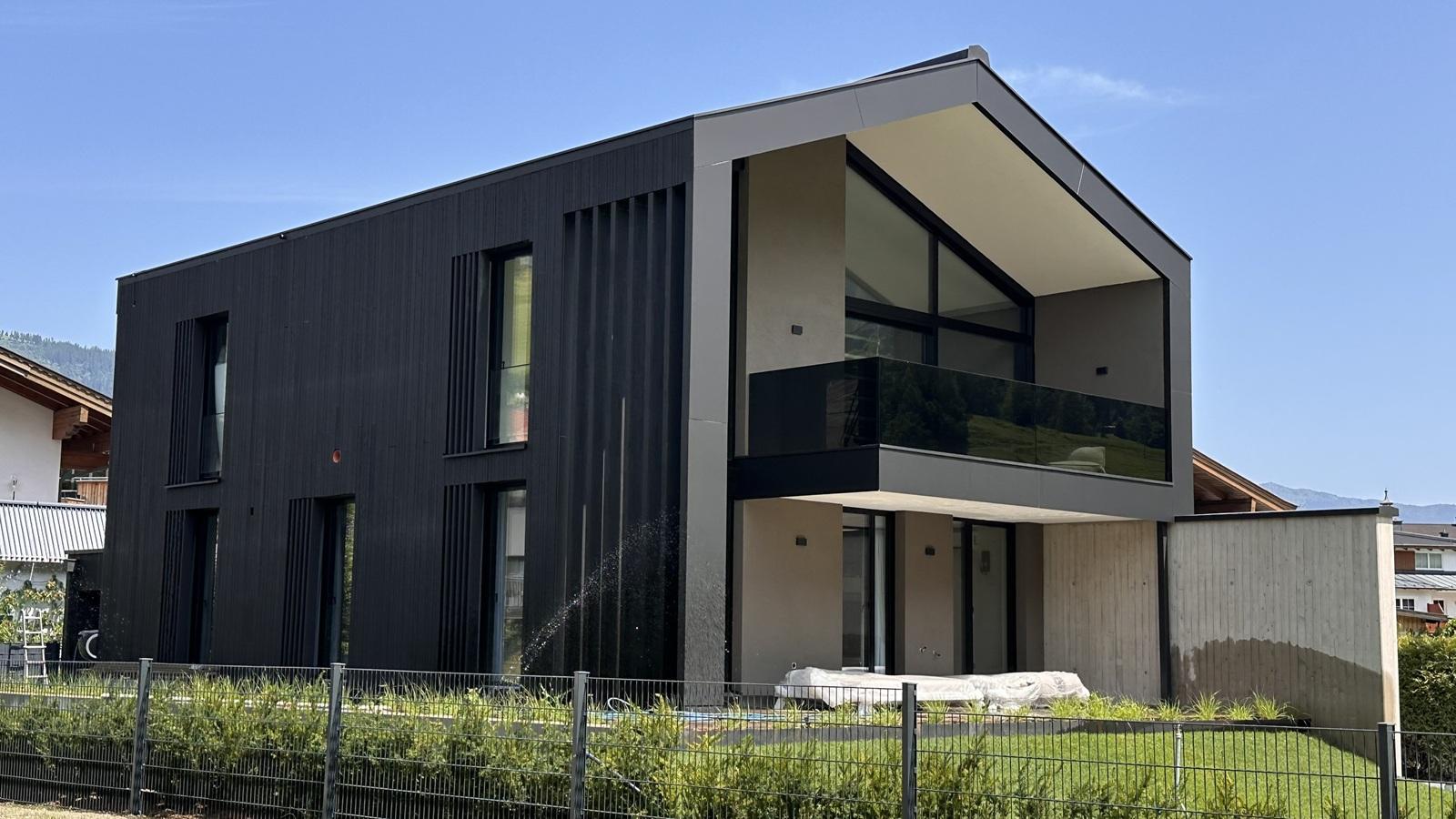 A modern house with a striking black facade and large windows. The balcony on the upper floor offers a view of the surroundings.