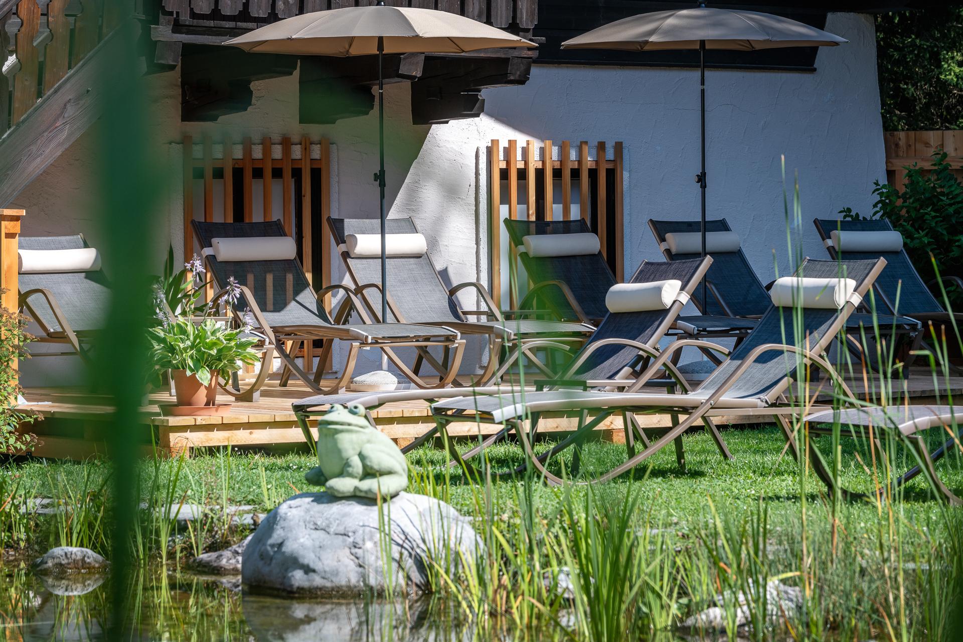 A relaxing terrace with lounge chairs and sun umbrellas. A frog sits on a stone in the greenery next to a pond.