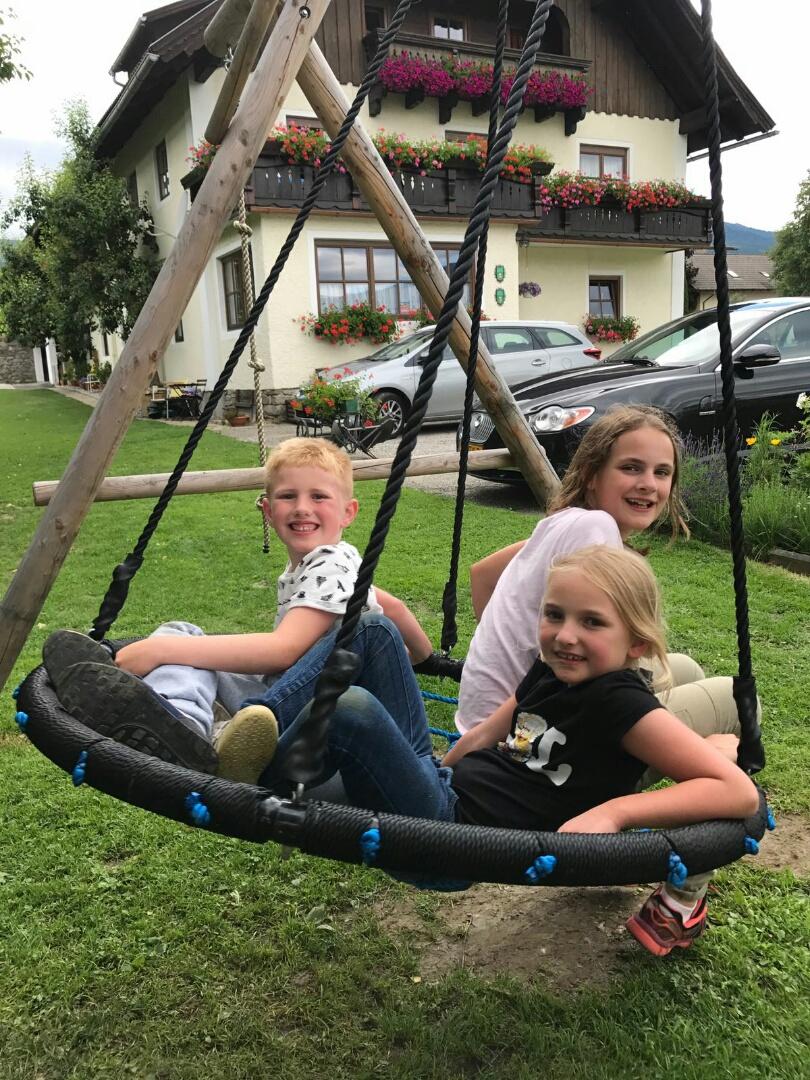 Three children are sitting on a large nest swing outdoors. In the background, a house with flower balconies and cars can be seen.