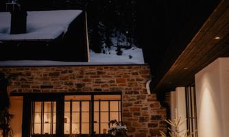 A cozy house in a snowy landscape at night. The trees and mountains in the background create a serene winter atmosphere.