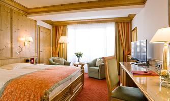 A cozy hotel room with wooden walls and a large window. It features a double bed, seating areas, and a television.