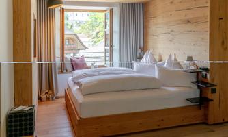 A modern hotel room with a large bed and wooden details. The window lets in a lot of light and offers a view of the surroundings.
