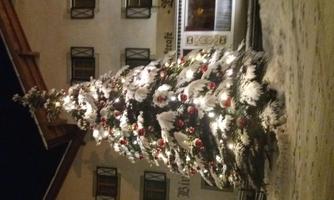 A festively decorated Christmas tree with lights and ornaments stands in front of a traditional building. The ground is covered with snow, creating a wintry atmosphere.