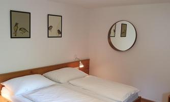 A simple bedroom with a double bed and two nightstands. Pictures of birds hang on the wall and a round mirror is also visible.
