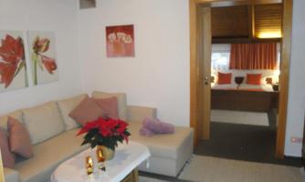 A cozy living space with a bright couch and decorative pillows. In the background, a bedroom is visible, warmly lit.