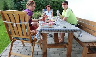 Three people are sitting at an outdoor table enjoying food and drinks. The table is covered with a checkered tablecloth and surrounded by greenery.