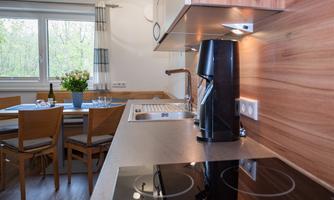 A modern kitchen with a stove, a coffee machine, and a sink. In the background, there is a dining area with wooden chairs and a table.