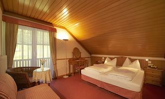 A cozy room with a double bed and wooden walls. There is a small table, a chair, and plenty of daylight coming through the windows.