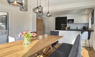 A modern dining area with a large wooden table and comfortable chairs. The stylish kitchen in the background is well-equipped and brightly lit.