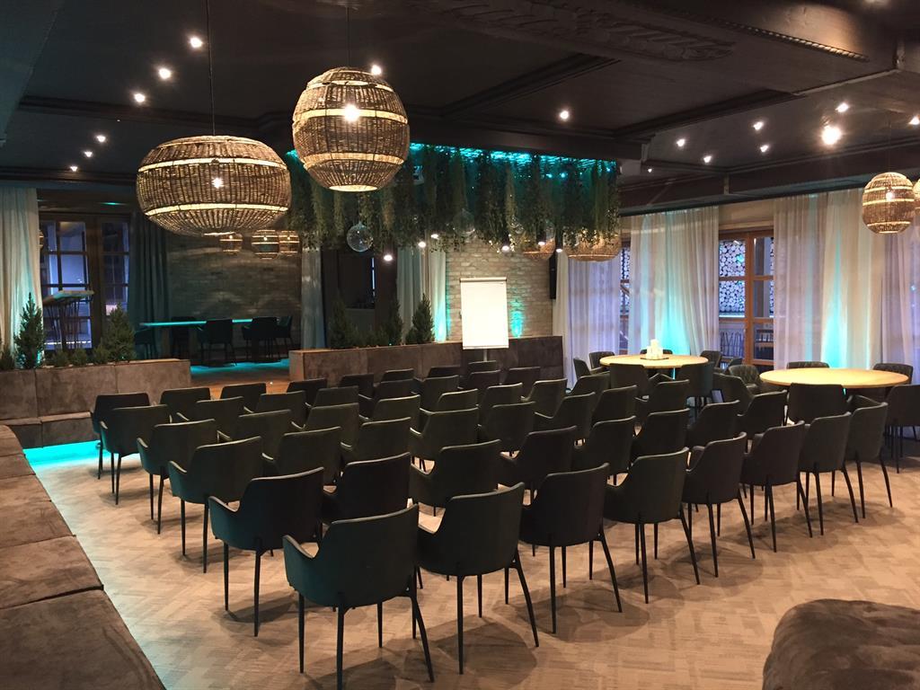 A modern conference room with rows of chairs and large hanging lights. The atmosphere is friendly and inviting, with soft lighting and plants.