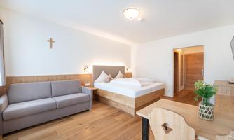 A modern room with a double bed, a sofa, and wooden furniture. The walls are light-colored and there is natural light coming through a window.