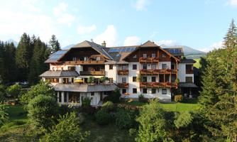 A cozy hotel surrounded by trees and a green garden. There are balconies with plants and a sunny atmosphere.