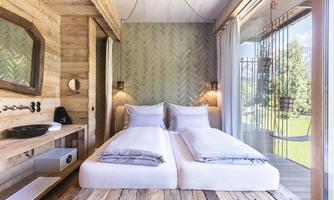 A modern bedroom with two single beds and rustic wooden walls. Large windows offer a view of the green surroundings.