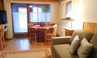 A cozy living room with a sofa, a dining table, and a television. Large windows with curtains and a warm wood design.