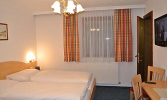 A cozy hotel room with two beds, a table, and chairs. The windows are adorned with checkered curtains, and there is a television on the wall.