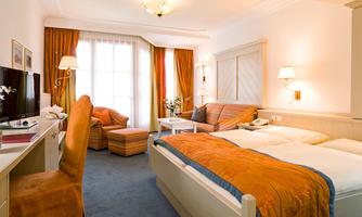 A cozy hotel room with two beds and bright decor. The room features a television and a sofa, as well as large windows with daylight.