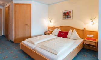 A cozy hotel room with a double bed and light walls. The room is simply furnished and has a warm, welcoming character.
