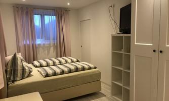 A modern bedroom with a cozy bed and a television. The walls are bright, and large windows provide plenty of daylight.