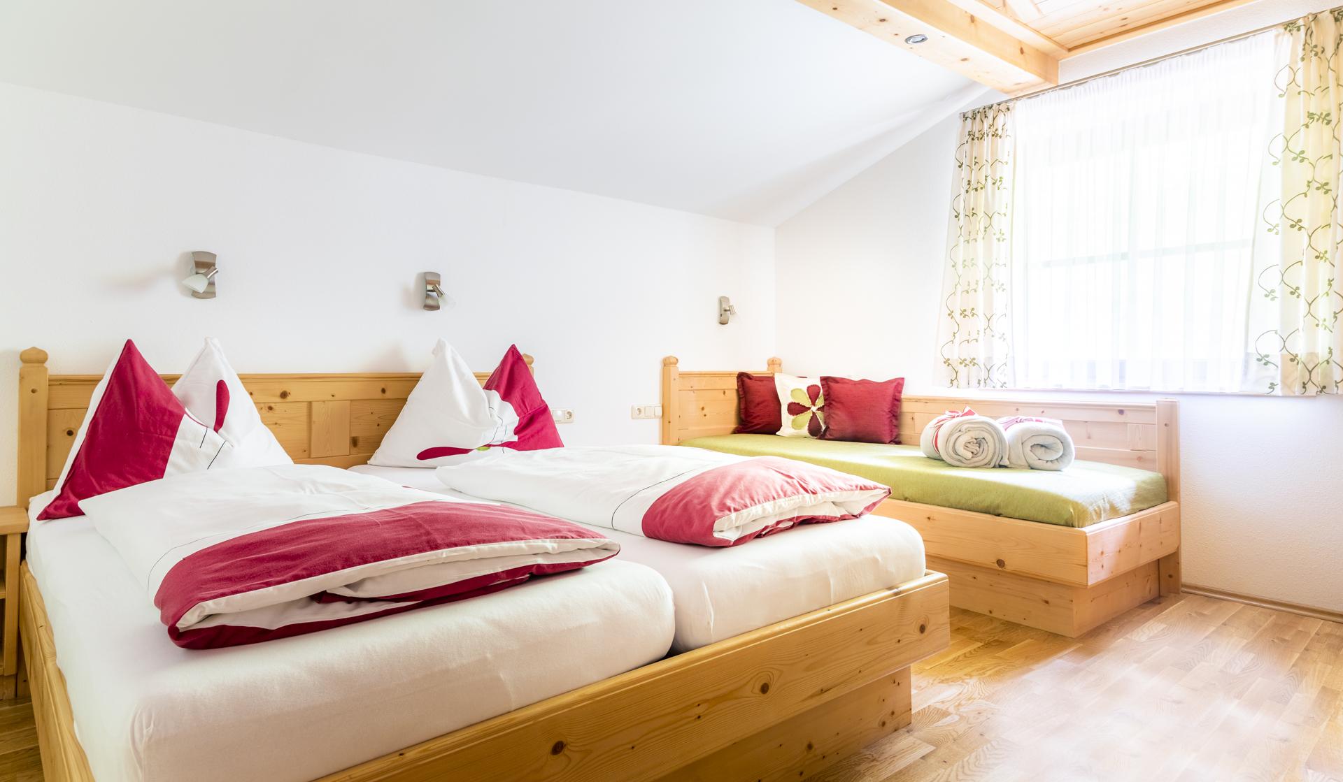 A bright room with two wooden beds. Decorated with red cushions and a window that lets in daylight.