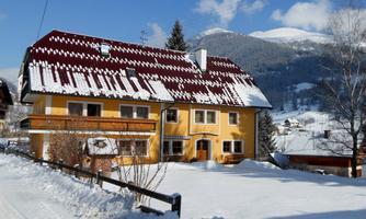 A yellow house with a red roof stands in the snow. In the background, snow-covered mountains and a blue sky can be seen.