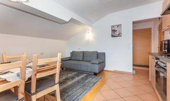 A cozy living kitchen with a dining table, chairs, and a gray sofa. The room has a bright and inviting atmosphere.