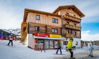 A wooden building in a ski resort with a layer of snow. Skiers are standing near the ski rental.