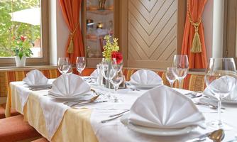 An elegantly set table with white napkins and glasses. The room has large windows and is brightly decorated.